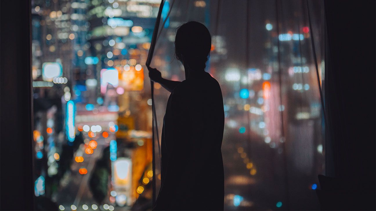 Silhouette of a person in hotel room at night looking out window at bright city lights